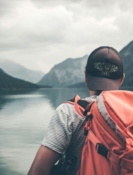 man with red hiking backpack facing body of water and mountains at daytime